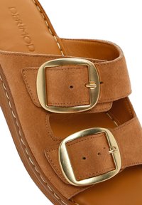 Bruine suède slidesandalen met twee goudkleurige gespen, een comfortabele voetbed en witte stiksels langs de rand. Vlakke zoolontwerpen.