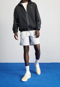 Man i svart zip-up jacka, ljusa shorts, vita strumpor och beigefärgade Adidas-sneakers som står på blått golv mot vit vägg.