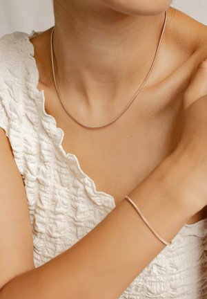 Silberne Halskette mit einem glatten, abgerundeten Design, die auf dem Schlüsselbein ruht, kombiniert mit einem schlanken Armband am Handgelenk, beides vor einem strukturierten weißen Oberteil.