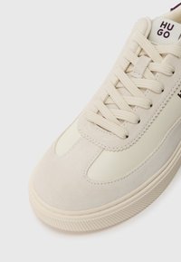 Αθλητικό παπούτσι με μπεζ σουέτ και καμβά στο πάνω μέρος, διαθέτοντας λευκές κορδόνες, υφασμάτινη σόλα και λογότυπο στη γλώσσα. Κλα�σικός χαμηλός σχεδιασμός.