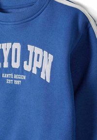 Modra trenirka z belim vezenim napisom "TOKYO JPN" in "KANTÓ REGIJA EST 1997." Ima rebrasto ovratnico in bele ramenske akcenta.