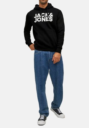 Man draagt een zwarte hoodie met de tekst "JACK & JONES", blauwe relaxed-fit jeans en zwart-witte sneakers, staand tegen een effen achtergrond.