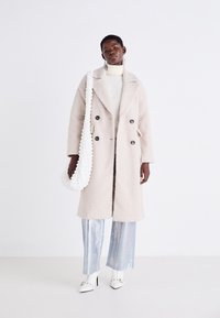 Cappotto doppiopetto rosa chiaro in tessuto testurizzato, abbinato a pantaloni larghi argentati lucidi, scarpe con tacco bianche e una borsa bianca.