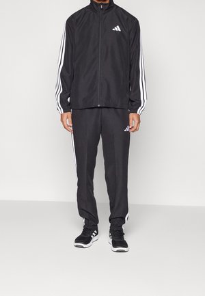 Homme portant un survêtement Adidas noir avec des bandes blanches sur les manches et les jambes, debout devant un fond clair uni.