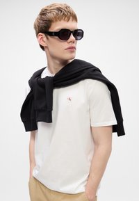 T-shirt branco de mangas curtas com um pequeno logótipo vermelho, acompanhado de um suéter preto colocado sobre os ombros e óculos de sol octogonais pretos.