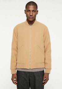 Beige Bomberjacke mit Reißverschluss vorne, Rippbündchen und seitlichen Taschen. Hergestellt aus weichem Stoff, mit Rundhalsausschnitt und strukturiertem Design darunter.