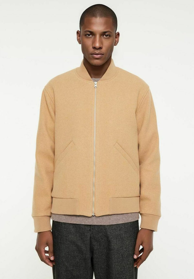 Beige Bomberjacke mit Reißverschluss vorne, Rippbündchen und seitlichen Taschen. Hergestellt aus weichem Stoff, mit Rundhalsausschnitt und strukturiertem Design darunter.