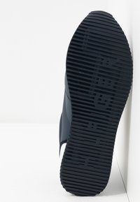 Czarna gumowa podeszwa buta o teksturowanej powierzchni, z poziomymi i pionowymi rowkami, lekko zwężającym się kształcie oraz wytłoczonym logo.