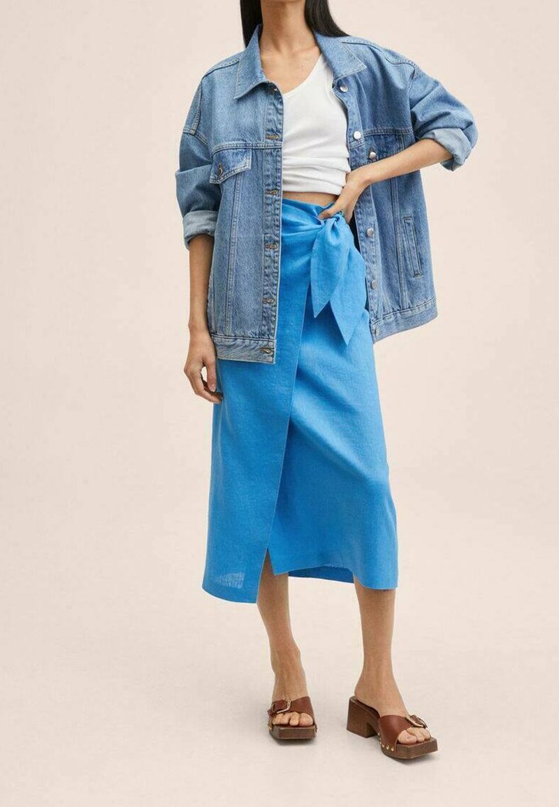 Giacca di jeans con le maniche arrotolate, crop top bianco e gonnellina in lino blu legata in vita. Sandali con piattaforma marroni.