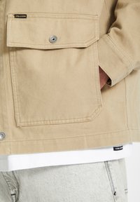 Chaqueta beige hecha de tejido texturizado, con un gran bolsillo en el pecho, cierre de botón a presión de metal y etiqueta de marca Völcom en el dobladillo.