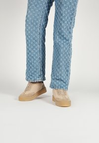 Beige sneakers with a textured, fuzzy upper and light brown suede accents, paired with blue, patterned, loosely fitting denim pants.