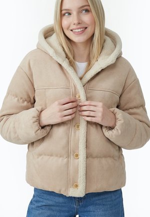 Junge Frau, die Knöpfe an einer beigen, sherpa-gefütterten Kapuzenjacke schließt, trägt blaue Jeans, lächelt leicht, vor weißem Hintergrund.