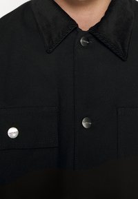 Primo piano di una giacca nera con colletto in corduroy, due tasche sul petto con bottone e tre bottoni a pressione metallici centrali.