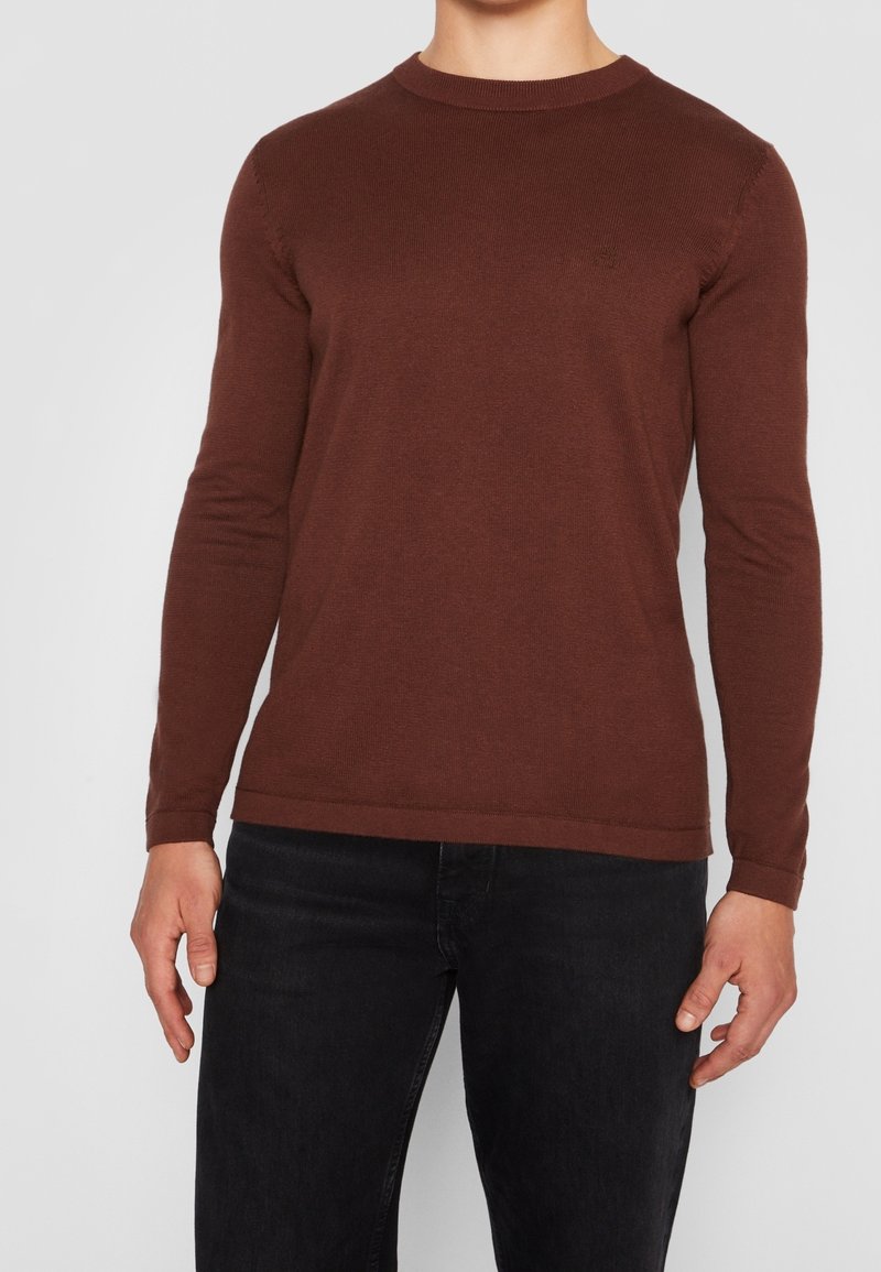 Pull brun à manches longues en matériau doux, avec un col rond et une coupe ajustée, présentant un détail de logo subtil sur la poitrine.