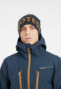 Chaqueta impermeable azul marino con cremalleras y logotipo en color beige; cuenta con un alto cuello y capucha, complementada con un gorro de punto a juego con "PROTEST" en naranja.