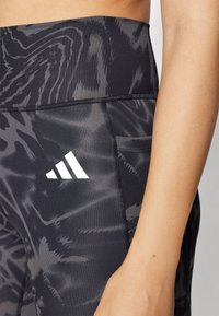 Leggings neri con un motivo testurizzato in tonalità di grigio, con il logo bianco di Adidas sulla coscia sinistra e dettagli di cucitura distintivi.