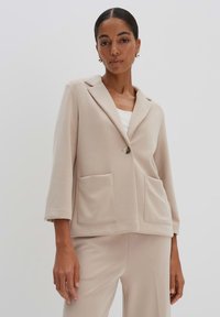 Blazer beige in tessuto morbido, con colletto a punta, chiusura con un bottone e due grandi tasche frontali. Vestibilità sartoriale con maniche a tre quarti.