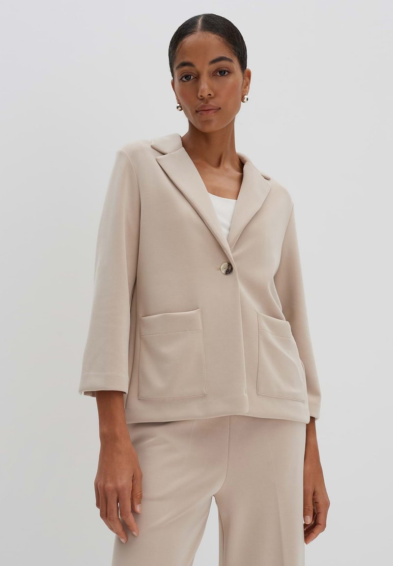 Blazer beige in tessuto morbido, con colletto a punta, chiusura con un bottone e due grandi tasche frontali. Vestibilità sartoriale con maniche a tre quarti.