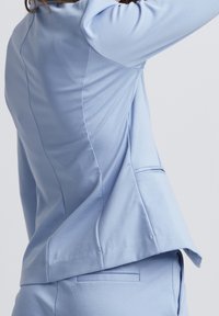 Ljusblå blazer med en slät yta, som har en något utfälld silhuett, subtila sömdetaljer och en enda sydd ficka på baksidan.