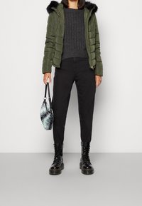 Doudoune vert olive avec une capuche doublée de fourrure noire, associée à un pull côtelé gris foncé, un pantalon noir fuselé et des bottines noires à lacets.