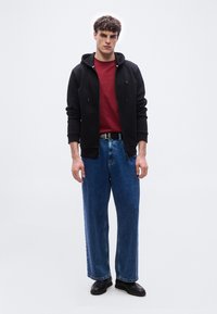 Νεαρός άνδρας φορώντας μαύρο φούτερ με φερμουάρ, κόκκινο πουκάμισο, μπλε παντελόνι με φαρδύ πόδι, μαύρη ζώνη και μαύρα παπούτσια, στέκεται μπροστά σε απλό φόντο.