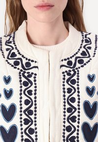 Hvid tekstureret stof cardigan med blå og marineblå broderi af hjerter og mønstre langs fronten og halsudskæringen, med en simpel lukning.