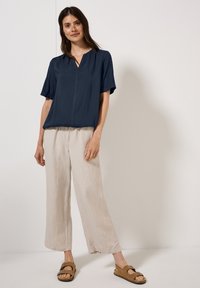 Blusa navy a maniche corte con scollatura a V, abbinata a pantaloni beige chiaro a gamba larga e sandali marroni. Tessuto morbido, vestibilità rilassata.
