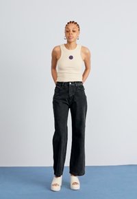 Débardeur beige avec un logo circulaire violet, associé à un jean noir évasé taille haute et des sandales à plateforme blanc cassé sur un fond bleu.