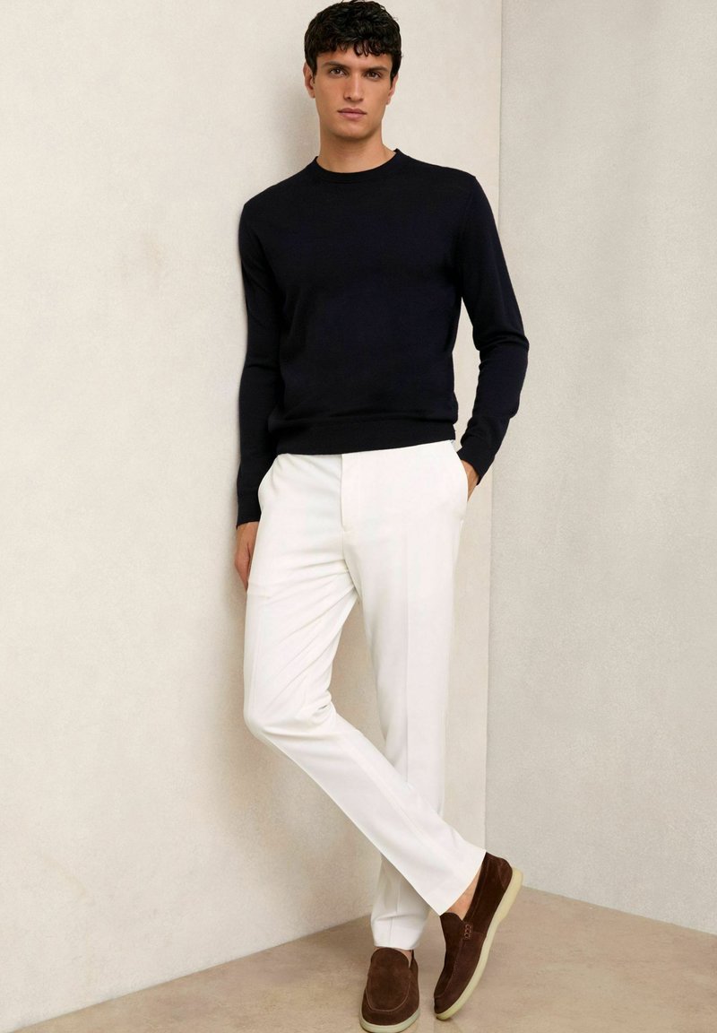 Mørkeblå strikket sweater, hvide skræddersyede bukser, brune ruskinds slip-on sko. Model står mod en lys neutral baggrund med hænderne i lommerne.