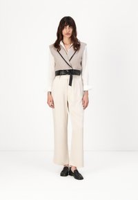 Gilet en mélange de laine beige avec une ceinture en cuir noir, assorti à un pantalon large crème et des mocassins noirs, sur fond blanc uni.