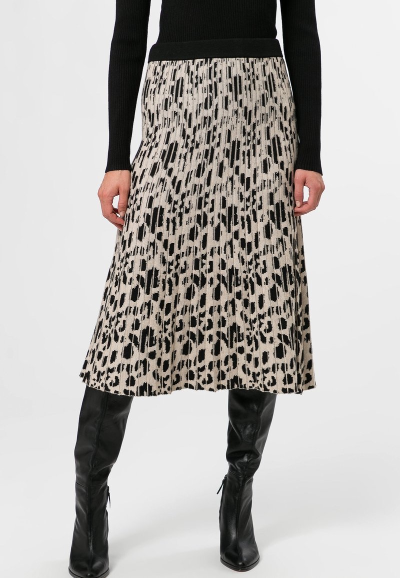Femme portant une jupe midi plissée imprimé léopard noir et beige, associée à des bottes en cuir noires montantes jusqu’aux genoux et un pull noir côtelé.