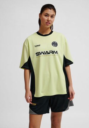 Młoda kobieta w żółto-czarnym sportowym trykocie i czarnych szortach, stojąca na jednolitym szarym tle.