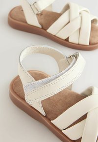 Vita konstläder sandaler med korslagda remmar, texturerad fotbädd och brun gummisul. Silverdetalj på remmen ger extra detalj.