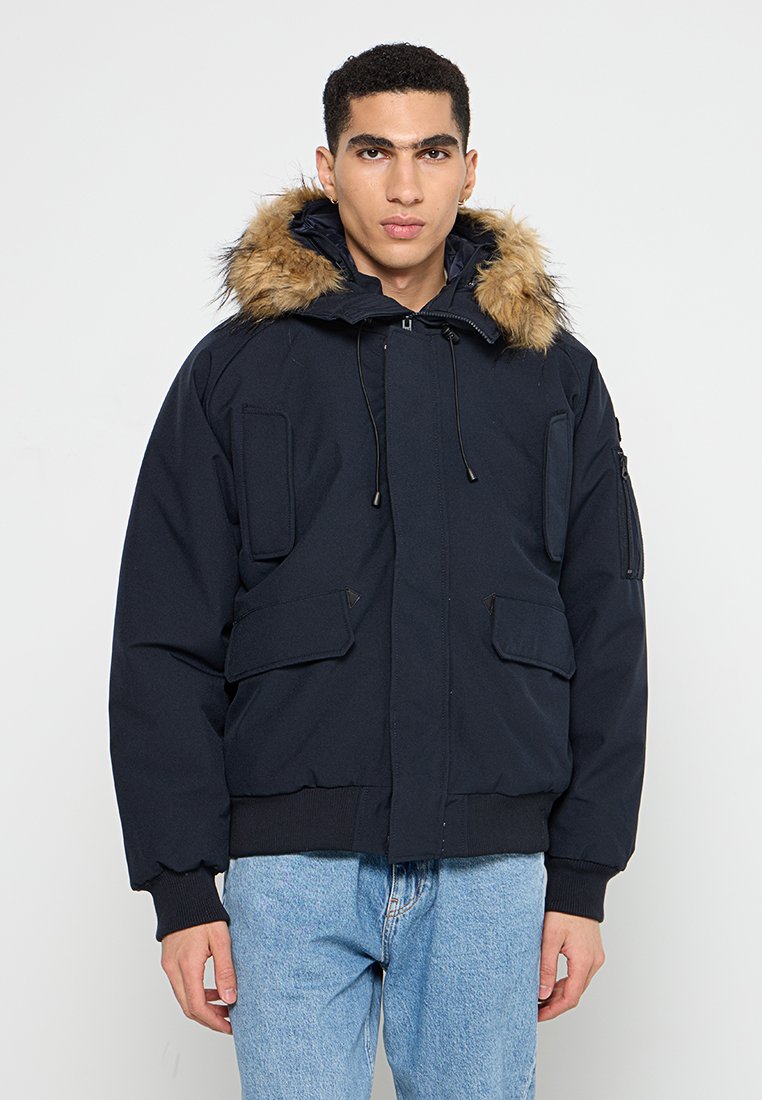 Jeune homme portant une veste d'hiver bleu marine avec capuche en fourrure et jeans bleu clair, se tenant devant un fond blanc.