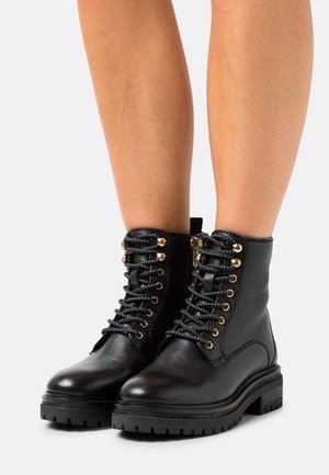 Zwarte leren enkellaarsjes met dikke profielzolen, zwarte veters door goudkleurige oogjes geregen, gedragen door een persoon met blote benen op een witte achtergrond.