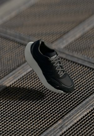 Baskets noires avec semelle blanche et lacets gris flottant au-dessus d'un sol en grille métallique texturée.