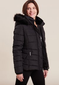 Donna sorridente che indossa un giubbotto invernale nero trapuntato con cappuccio bordato di pelliccia, chiuso con zip e bottoni, in piedi su uno sfondo neutro.