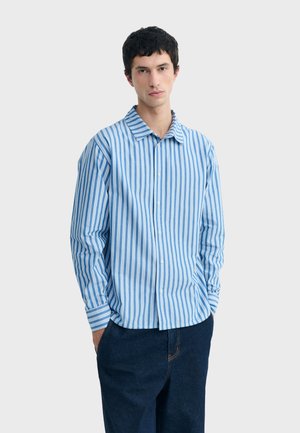 Junger Mann trägt ein blau-weiß vertikal gestreiftes Hemd mit Knöpfen und dunkelblaue Hose, steht mit den Händen in den Taschen vor einem schlichten Hintergrund.