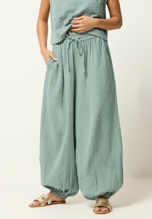 Frau trägt lockere, hellgrüne Hose mit Kordelzug und elastischen Bündchen sowie goldene flache Sandalen, eine Hand in der Tasche und die andere justiert das Oberteil.