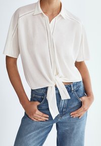 Weißes kurzärmliges Hemd mit Knopfleiste und Bindegürtel, das subtile Nähte aufweist. Getragen mit hoch taillierten blauen Jeans.