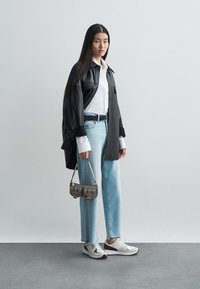Svart oversized skjorte, hvit skjorte med knapper, lyseblå jeans og hvite joggesko med røde detaljer. Liten teksturert veske holdt i den ene hånden.