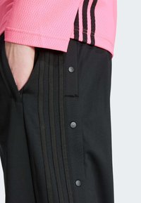 Sorte bukser med trykknapper og lodrette striber ved siden af en lys pink mesh-top med sorte detaljer og en tekstureret overflade.