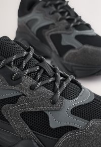 Baskets noires et grises comprenant du mesh, du daim et des matériaux synthétiques. Le design inclut des accents texturés et des motifs de couture contrastés.