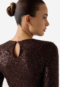 Top marrone con paillettes, scollo rotondo e apertura a goccia sul retro. Modella con orecchini pendenti oro. Capelli lisci raccolti in uno chignon.