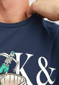 Camiseta de algodón azul marino con un gráfico de un esqueleto en pantalones cortos verdes y una gorra, con letras blancas que dicen "K &" de forma destacada.