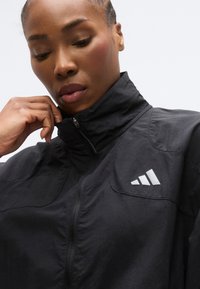 Must musta lukuga jakk kõrge kraega, millel on tekstuurne kangas ja rinnal peegeldav Adidas logo, loodud aktiivseks kandmiseks.