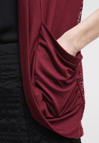 Main reposant dans une grande poche en tissu bordeaux sur un vêtement avec de la dentelle et un tissu noir texturé visible.