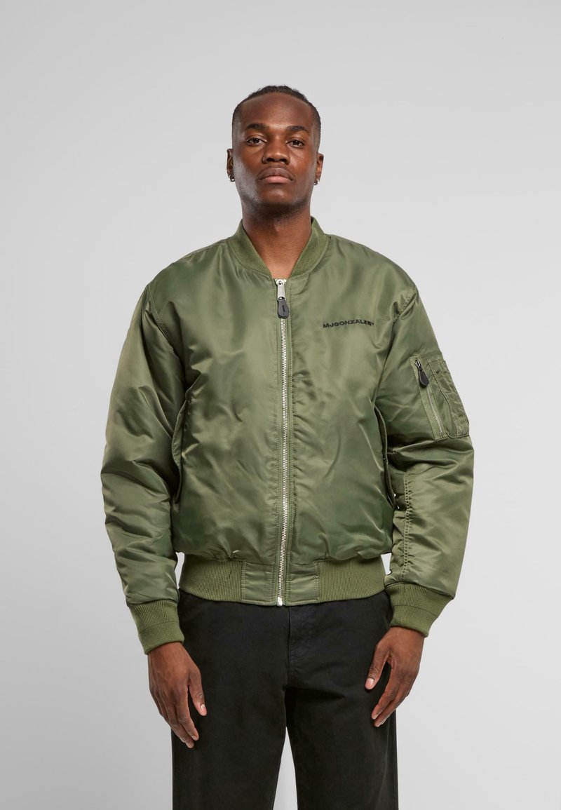Giacca bomber in satin verde oliva con chiusura a zip, polsini e orlo a coste. Presenta una tasca laterale con zip e un logo nero sul petto.