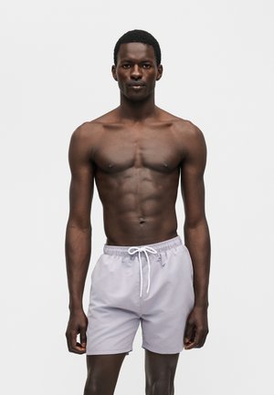 Homme athlétique à la peau foncée debout sur un fond uni, portant un short de bain gris clair avec un cordon blanc, regardant devant lui.