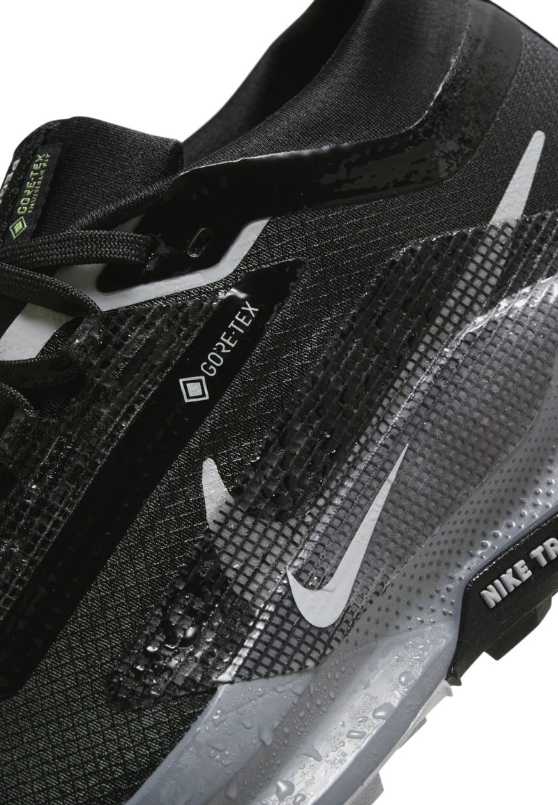 Zapatillas negras de GORE-TEX con parte superior texturizada y acabado brillante, detalles en gris y un logo de swoosh blanco en el lateral. Se observan gotas de agua.